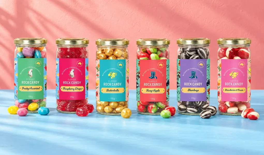 Six jars of Botanical Confectionery Co. classic rock candy, from Fruity Assorted to Strawberries & Cream, with loose sweets in front