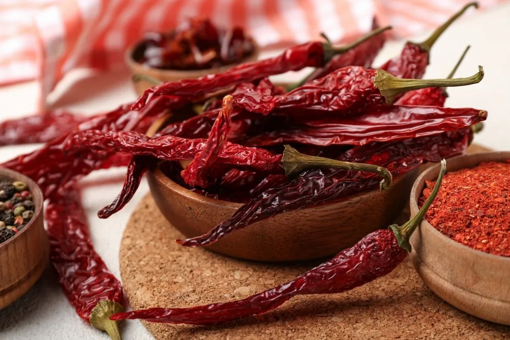 Dried red chipotle chillies in a wooden bowl, with bowls of chilli flakes and mixed peppercorns