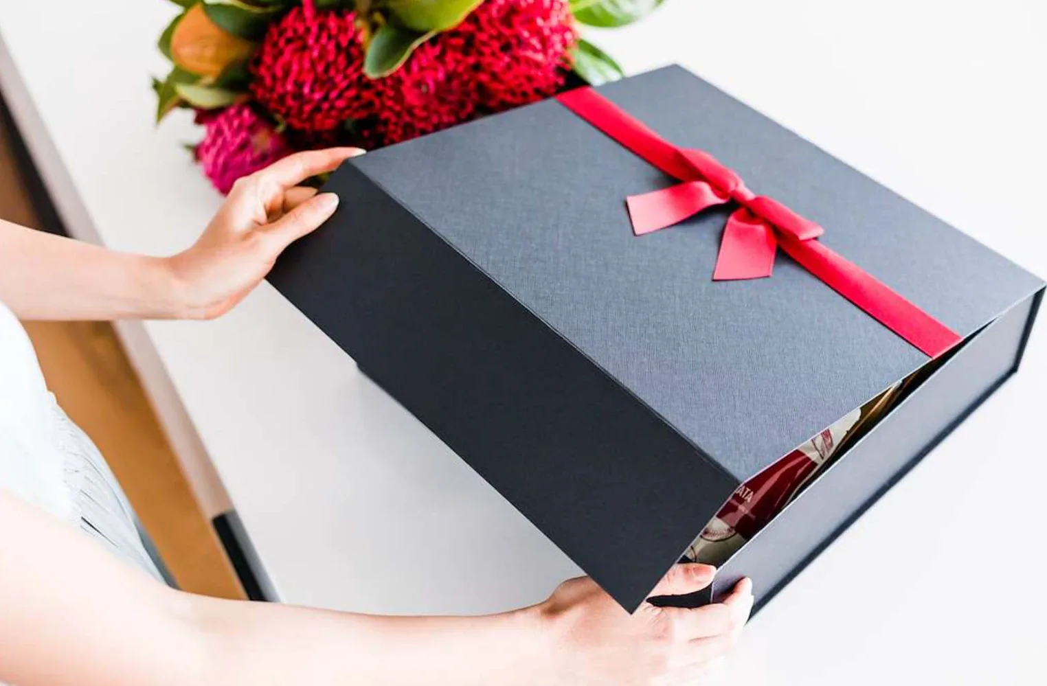 Hands opening a charcoal magnetic-lid hamper box tied with red satin ribbon, gourmet products inside