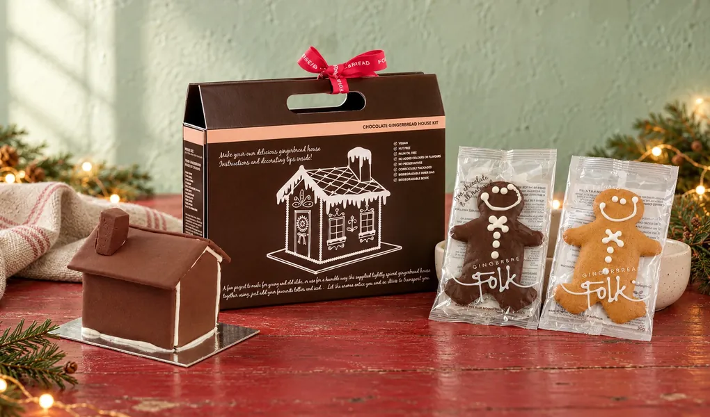 Gingerbread Folk chocolate gingerbread house kit with a built house and two wrapped gingerbread people on a red Christmas table