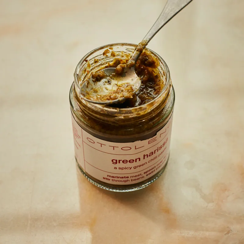 An open jar of Ottolenghi green harissa with a spoon resting in the chunky green chilli paste, on a pale marble surface