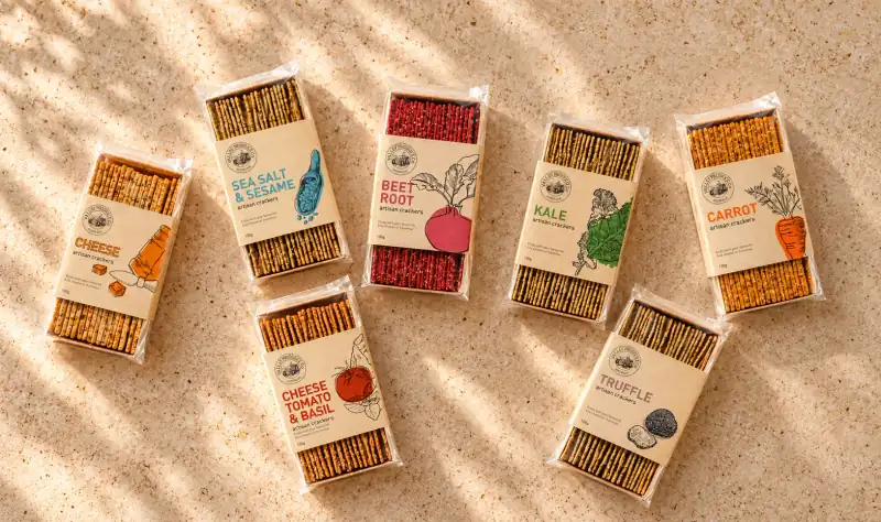 Seven packs of Valley Produce Company artisan crackers in cheese, sea salt and sesame, beetroot, kale, carrot, cheese tomato and basil, and truffle flavours on a sunlit stone bench